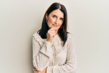 Orta yaşlı esmer kadın, elleri çenesinde gündelik kıyafetler giyip soru soruyor, düşünceli bir yüz ifadesi var. Düşünceli bir yüzle gülümsüyor. şüphe kavramı. 