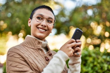 Kısa saçlı genç İspanyol kadın akıllı telefon kullanarak mutlu bir şekilde gülümsüyor.