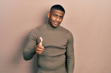 Balıkçı yaka kazak giyen genç siyahi bir adam mutlu bir el hareketi yapıyor. Kameraya bakıp başarıyı gösteren ifade. 