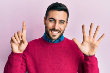 Latin asıllı genç adam gündelik giysiler giyip altı numaralı parmaklarıyla işaret ederken kendinden emin ve mutlu bir şekilde gülümsüyor.. 