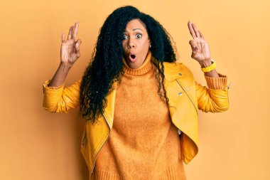 Orta yaşlı Afro-Amerikalı kadın yün kazak ve deri ceket giyiyor. Şaşırmış ve şaşırmış görünüyor. Tamam sembolünü parmaklarıyla yapıyor. çılgın ifade 