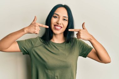 Gündelik tişört giyen, neşeli gülümseyen, parmaklarıyla ve ağzıyla işaret eden genç İspanyol kız. diş sağlığı kavramı. 