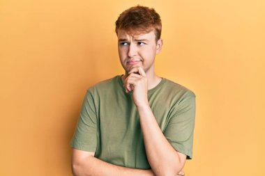 Gündelik yeşil t-shirt giyen genç beyaz adam şüpheye odaklanmış, çenesinde parmak olan ve kafasını kaldırıp merak ediyor. 