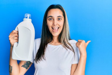Deterjan şişesini tutan genç İspanyol kadın baş parmağını yukarı kaldırıyor ve ağzı açık gülümsüyor. 