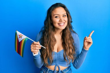 Genç İspanyol kız zimbabwe bayrağı tutuyor gülümsüyor mutlu bir şekilde el ve parmak kenarı işaret ediyor. 