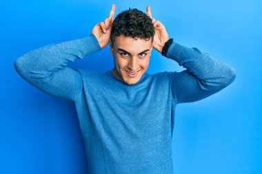 İspanyol genç adam günlük kazak giyiyor komik ve çılgın poz veriyor parmakları tavşan kulağı gibi, neşeli gülümsüyor 