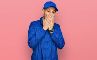 Kel ve sakallı bir adam, inşaatçı üniforması giyiyor. Gülüyor ve utanıyor. Ağzı ellerle, dedikoduyla ve skandal konseptiyle örtüyor. 