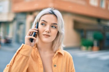 Rahatlamış yüzlü genç sarışın kız şehirde akıllı telefondan konuşuyor..