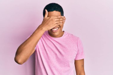 Genç, yakışıklı, pembe tişört giyen, gülümseyen ve elleri yüzünde, şaşırtmak için gözlerini kapatan bir İspanyol. kör kavram. 