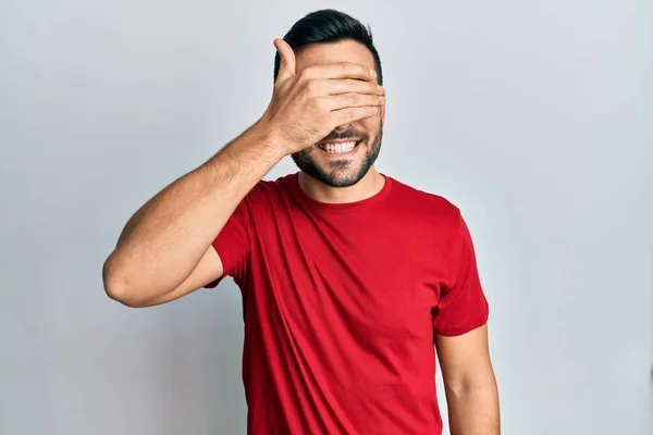カジュアルな服を着た若いヒスパニック系の男が笑顔で 驚きのために目をカバー顔に手で笑っています 視覚障害の概念 — ストック写真