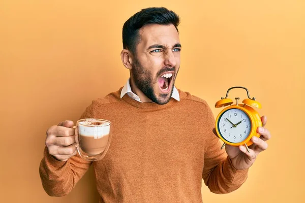 Young hispanic man holding coffee and alarm clock angry and mad screaming frustrated and furious, shouting with anger. rage and aggressive concept.