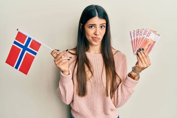 拿着诺威国旗和克朗钞票的年轻惊慌失措的女人表示怀疑和紧张 因为问题皱着眉头 消极的人 — 图库照片