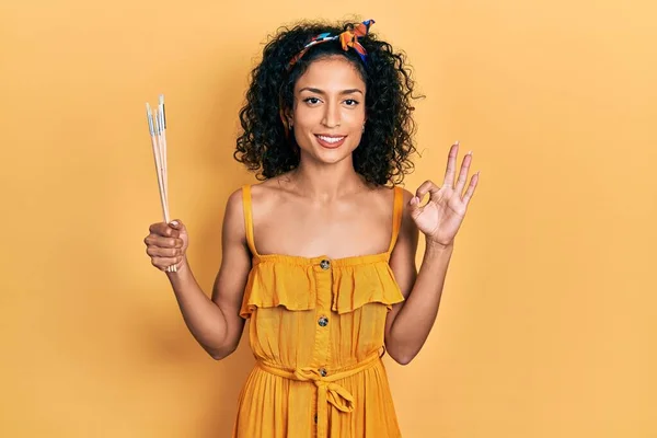Young Latin Girl Holding Paintbrushes Doing Sign Fingers Smiling Friendly — Stock Photo, Image