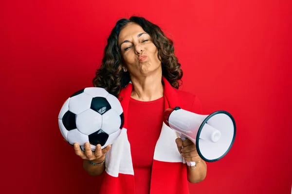 Middle age hispanic woman football hooligan holding ball and using megaphone looking at the camera blowing a kiss being lovely and sexy. love expression.