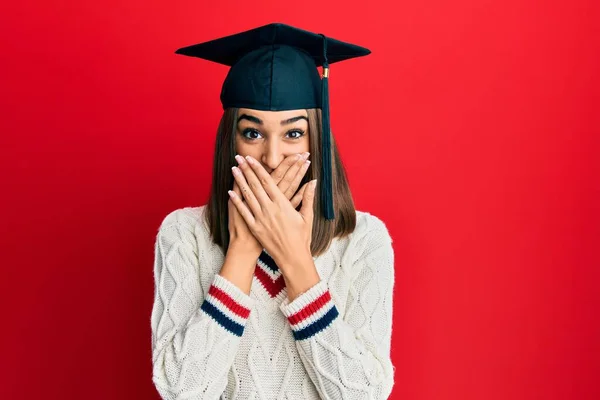 卒業キャップを被った若いブルネットの女の子は間違いのために手で口を覆いショックを受けた 秘密の概念 — ストック写真