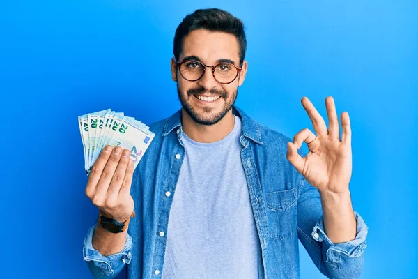 Joven Hombre Hispano Sosteniendo Montón Billetes Euros Haciendo Bien Signo —  Fotos de Stock