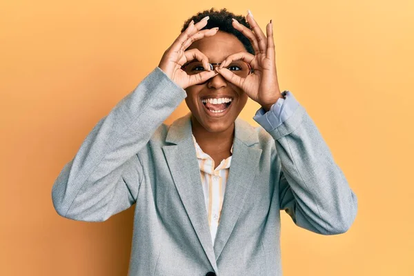 年轻的非洲裔美国女孩穿着商务夹克 戴着眼镜 像双筒望远镜一样做手势 伸出舌头 眼睛透过手指看着 疯狂的表达 — 图库照片