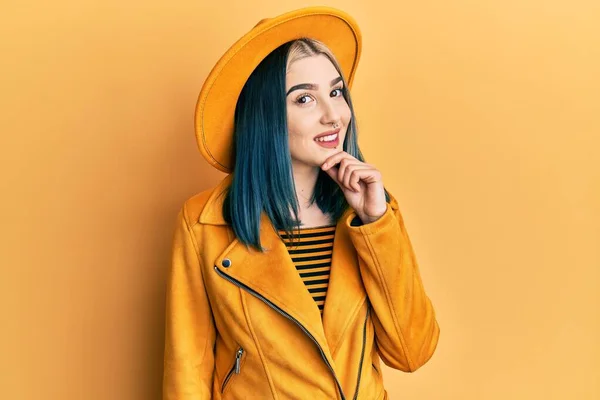 黄色の帽子と革のジャケットを身に着けている若い現代の女の子は カメラを見て自信を持って交差腕と手であごに提起笑みを浮かべている ポジティブな — ストック写真