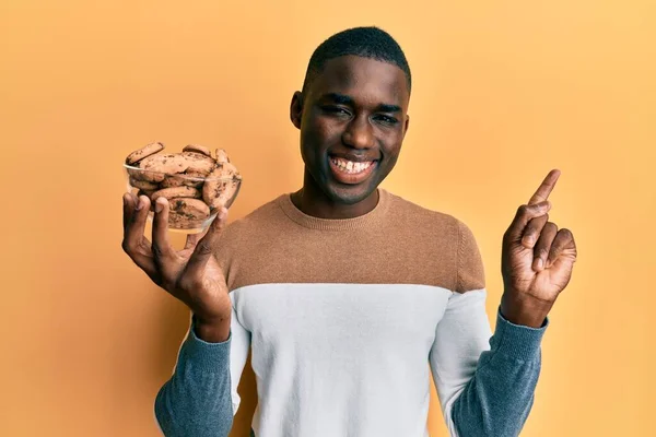 年轻的非洲裔美国人拿着装有巧克力片饼干的碗 高兴地微笑着 手指手画脚地指向旁边 — 图库照片