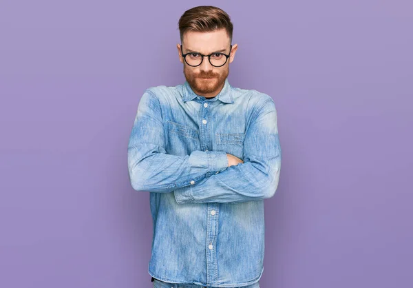 Young redhead man wearing casual denim shirt skeptic and nervous, disapproving expression on face with crossed arms. negative person.