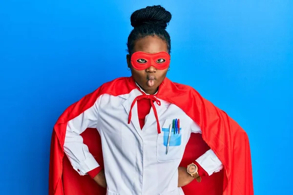 Young african american woman wearing doctor uniform and super hero costume making fish face with mouth and squinting eyes, crazy and comical.