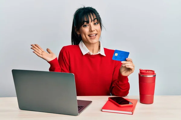 Young brunette woman with bangs working at the office using laptop and credit card celebrating achievement with happy smile and winner expression with raised hand