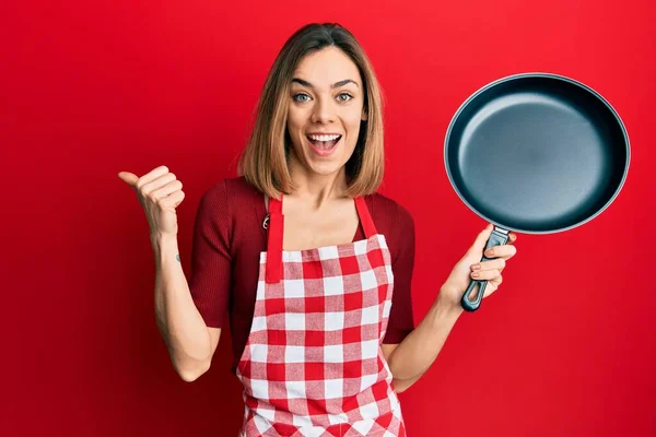 年轻的高加索金发女人穿着厨师围裙 手拿着锅子 手指指向旁边 笑容满面 张开嘴 — 图库照片