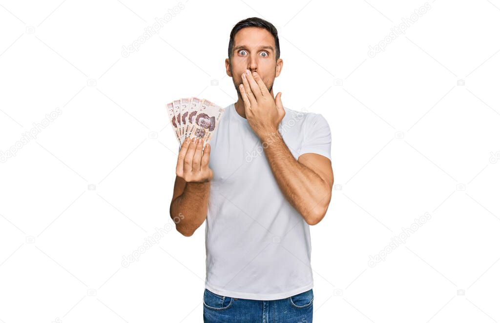 Handsome man with beard holding 50 mexican pesos banknotes covering mouth with hand, shocked and afraid for mistake. surprised expression 