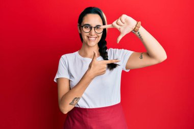 Genç İspanyol kadın profesyonel garson önlüğü giyiyor gülümsüyor ve elleri ve parmakları gülen yüzüyle çerçeve yapıyor. yaratıcılık ve fotoğrafçılık kavramı. 