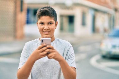 Sevimli Latin çocuk şehirde akıllı telefon kullanarak mutlu bir şekilde gülümsüyor..