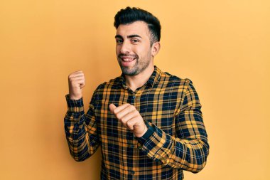 Gündelik kıyafetler giyen genç İspanyol adam arka tarafı işaret ediyor. Elleri havada, kendinden emin bir şekilde gülümsüyor. 