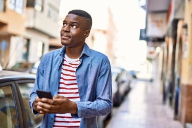 Genç Afrikalı Amerikalı adam şehirde akıllı telefon kullanarak mutlu bir şekilde gülümsüyor..