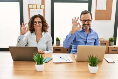 Orta yaşlı İspanyol bir kadın ve ofiste dizüstü bilgisayarı olan bir adam gülümsüyor ve el ve parmaklarıyla 