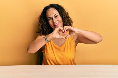 Güzel orta yaşlı kadın, gündelik giysiler giyip masaya oturmuş, aşıkken gülümsüyor, elleriyle kalp sembolü yapıyor. romantik kavram. 