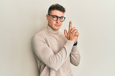 Balıkçı yaka kazak giyen İspanyol genç adam sembolik bir silah tutuyor, el hareketleriyle cinayet silahlarıyla oynuyor, kızgın suratlı. 