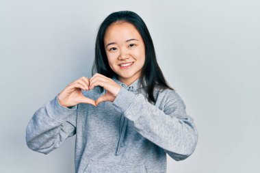 Gündelik kıyafetler giyen genç Çinli bir kız. Sevgiyle gülümsüyor. Kalp sembolü ve elleri ile şekil veriyor. romantik kavram. 