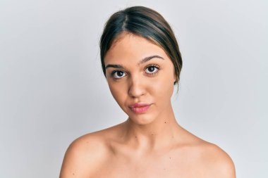 Genç esmer kadın üstsüz duruyor. Cildini gösteriyor. Düşünme tavrı sergiliyor. 