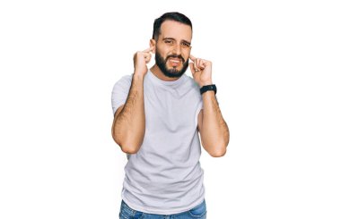 Gürültülü müzik sesi için sakallı, günlük beyaz tişörtlü, kulaklarını parmaklarıyla kapatan genç bir adam. sağır kavramı. 