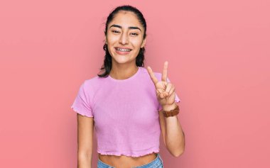 Dişlerinde diş teli olan İspanyol bir genç kız. Günlük kıyafetler giyiyor. İki numarayı gösteriyor ve gülümsüyor.. 