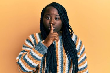 Saçları örgülü, kışlık süveter giyen genç siyahi bir kadın parmakları dudaklarında sessiz olmak istiyor. sessizlik ve gizli konsept. 