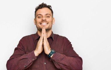 Gündelik kıyafetler giyen genç İspanyol adam elleriyle dua ediyor af diliyor kendinden emin bir şekilde gülümsüyor.. 