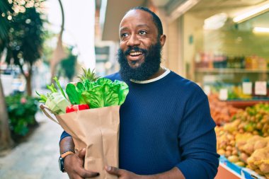 Sakallı Afrikalı Amerikalı bir adam elinde süpermarketten alınmış kağıt poşetler tutuyor.