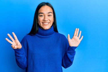 Genç Asyalı kadın, günlük kışlık süveter giyer ve sekiz numaralı parmaklarıyla gülümseyerek mutlu ve kendinden emin bir şekilde onu işaret eder.. 