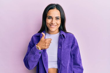 Genç esmer kadın gündelik giysiler giyip mutlu mesut el hareketi yapıyor. Kameraya bakıp başarıyı gösteren ifade. 