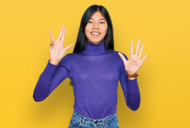 Güzel, genç Asyalı kadın gündelik kıyafetler giyip dokuz numaralı parmaklarıyla işaret ederken kendine güvenen ve mutlu bir şekilde gülümsüyor.. 