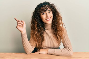 Latin asıllı genç bir kız, gündelik giysiler içinde masaya oturmuş gülümsüyor, mutlu bir şekilde elini ve parmağını kenarı gösteriyor. 