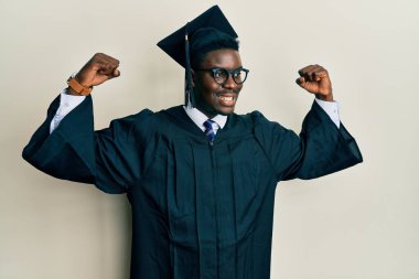 Mezuniyet şapkası ve tören cübbesi takan yakışıklı zenci gururla gülümseyen kol kaslarını gösteriyor. fitness konsepti. 