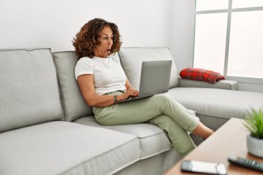 Orta yaşlı İspanyol kadın evde bilgisayar kullanarak çalışıyor. Şaşırmak için ağzı açık kalmış, korkmuş ve hayrete düşmüş. İnanmayan yüz. 