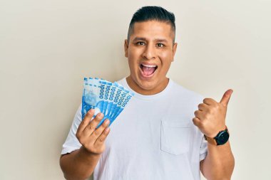 On bin Şili pezosu tutan genç latin adam başparmağıyla yan tarafı işaret ediyor, ağzı açık gülümsüyor. 