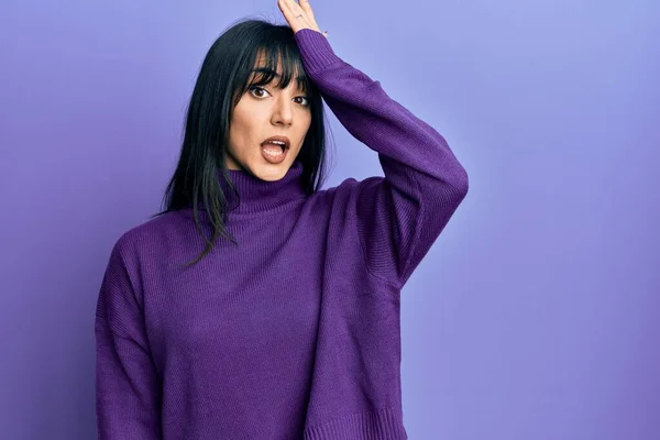 Young brunette woman with bangs wearing turtleneck sweater surprised with hand on head for mistake, remember error. forgot, bad memory concept.